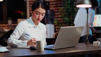 Asian worker sulking in office, having frown on face, feeling annoyed by having too much work to do. Employee writing down info on notepad, gloomily solving tasks late at night video