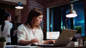 Businesswoman researching key data for company project, doing tasks in office overnight. Employee looking over accounting figures on laptop screen late at night, typing on keyboard, camera A video