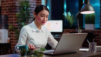 Businesswoman drinking cup of coffee while doing tasks in office overnight for company project. Employee enjoying hot beverage while looking over figures on laptop late at night, typing on keyboard video