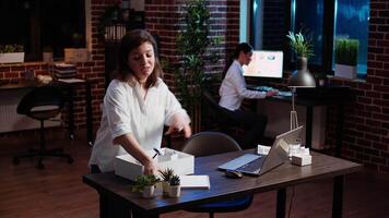 Woman packing office desk belongings, excited to go on holiday, hugging asian coworker. Joyful employee putting objects on workspace table in box, taking days off to go on vacation, camera A video