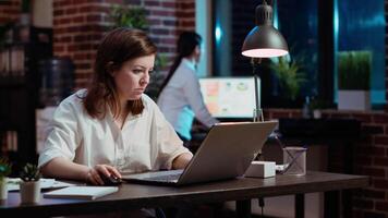 Office clerk looking through financial business documents on laptop late at night in workplace. Employee working on company project with deadline approaching soon during nightshift video