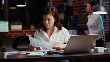 Worker looking over paperwork containing financial graphs, researching key data for company project. Businesswoman looking over accounting figures and charts on papers late at night at desk video