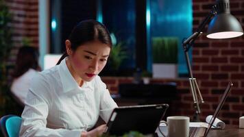 Businesswoman crosschecks data between financial charts and company internet website, doing tasks for team project in office overnight. Employee works at night, typing on laptop keyboard, camera B video