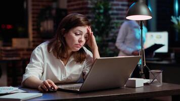 Businesswoman working overtime for company project, doing tasks in office overnight, struggling to remain awake. Tired employee yawning while looking over data on laptop screen late at night, camera B video