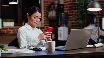 Smiling employee at work texting friends on cellphone, taking break from solving project tasks. Relaxed businesswoman using mobile phone to unwind in office late at night video