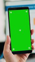 Vertical video Company worker holding smartphone with greenscreen template, using digital chromakey template on mobile gadget display. Employee presenting isolated mockup copyspace in business office.