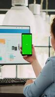 Vertical video Employee looks at greenscreen on phone, working on business operations at desk. Person holding smartphone device with isolated display showing mockup template, blank layout screen.