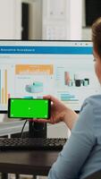 Vertical video Office worker looks at greenscreen on smartphone display app, analyzing isolated chromakey layout on mobile device at work. Woman holding phone that shows blank mockup template.