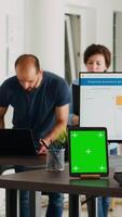 Vertikale Video leeren Schreibtisch mit grüner Bildschirm auf Tablette, modern Coworking Raum im kreativ Agentur Büro mit Angestellte beim Arbeit. leer Attrappe, Lehrmodell, Simulation isoliert Vorlage beim Arbeitsplatz, klein Geschäft Agentur.