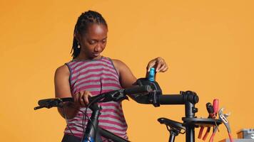 africain américain femme réglage en haut bicyclette réparation rester, en utilisant il à régler dérailleur, fixation cassé roues, studio Contexte. professionnel placement vélo sur support de travail à faire vérification sur il, caméra une video