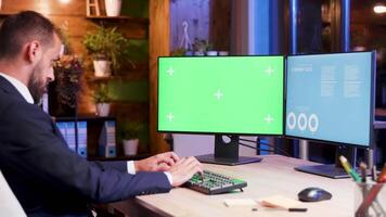 Businessman in suit typing on computer with green screen in his office with blue light from the moon coming from window. video