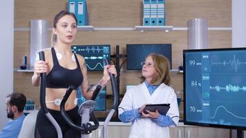 fêmea cientista monitoramento a coração atividade em fêmea atleta. fêmea atleta correndo com eletrodos em anexo dentro uma esporte teste Centro. video