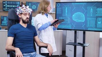 weiblich Neurologe lesen Daten von Gehirnwelle Scannen Headset auf pacient im ein Neurowissenschaften Labor. video