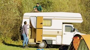 Boyfriend saying hello to her girlfriend who's relaxing on top of their vintage camper van. Couple relaxing in mountain wildnerss. video