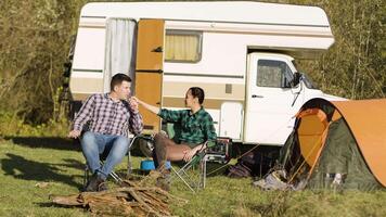 Hipster boyfriend kissing his girlfriend hand while sitting on camping seats in front of their vintage camper van. Relaxing Week-end video