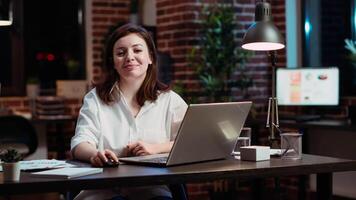 Portrait of smiling accountant researching key data for company project, doing tasks in office overnight. Happy employee sitting at desk in front of laptop, looking over accounting figures, camera B video