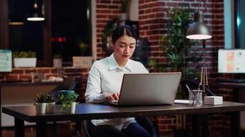 retrato de sonriente asiático mujer de negocios haciendo computadora Tareas para equipo proyecto en oficina. alegre empleado trabajando tarde a noche, mecanografía en ordenador portátil teclado siguiente a compañero de trabajo, cámara si video