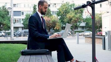 Young manager in suit working on the laptop outdoor in the city, slow motion. Next to him is an electric scooter video