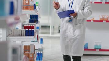 Druggist counting all pharmaceutical stock in pharmacy shop, working on inventory with checklist on clipboard. Pharmacist examining medical products on drugstore shelves. Camera 2. video