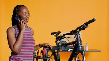 glad ingenjör på arbete tar ha sönder från reparation cykel till prata på telefon med vänner, fångst upp, studio bakgrund. leende mekaniker på smartphone ring upp med mates medan fixering cykel, kamera b video