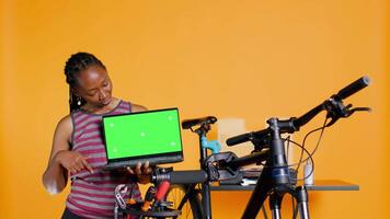 africain américain femme détient copie espace isolé écran ordinateur portable, en utilisant il à afficher bicyclette réparation boutique, studio Contexte. mécanicien promouvoir vélo fixation un service avec chrominance clé carnet video