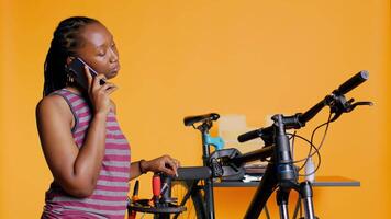 Afrikaanse Amerikaans vrouw pratend Aan telefoon met monteur, vragen voor helpen Aan repareren beschadigd fiets, studio achtergrond. bipoc persoon Aan telefoon telefoontje met fiets ingenieur, camera b video