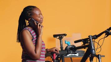 Afrikaanse Amerikaans vrouw pratend Aan telefoon met monteur, vragen voor helpen Aan repareren beschadigd fiets, studio achtergrond. bipoc persoon Aan telefoon telefoontje met fiets ingenieur, camera een video