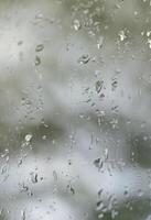 A photo of rain drops on the window glass with a blurred view of the blossoming green trees. Abstract image showing cloudy and rainy weather conditions