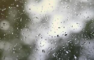 A photo of rain drops on the window glass with a blurred view of the blossoming green trees. Abstract image showing cloudy and rainy weather conditions