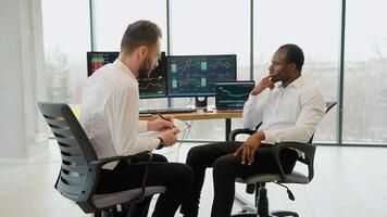 Two crypto traders sitting in front of computers, making professional analysis of candlestick chart, creating strategy, holding pen and notepad, looking at monitor video