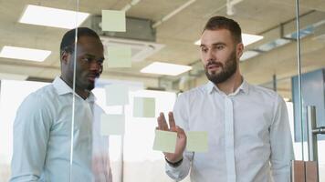 Two businessmen in meeting brainstorming and discussing post it notes stuck on glass wall at office video