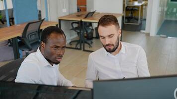 Two diverse traders sitting at desk at office together monitoring stocks data candle charts on screen video