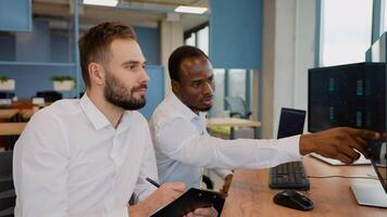 Two men traders sitting at desk at office together monitoring stocks data candle charts on screen, side view video