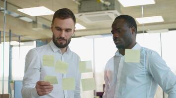 Two businessmen in meeting brainstorming and discussing post it notes stuck on glass wall at office video