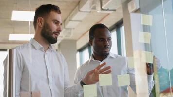 vielfältig Mannschaft von zwei Angestellte während Arbeiten zusammen während Brainstorming und Stehen hinter Glas Mauer mit klebrig bunt Papiere video