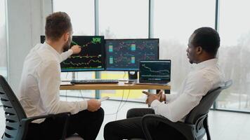 Two diverse colleagues traders talking to each other, looking at graphs while sitting in the office in front of multiple computer screens video