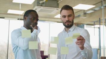 vielfältig Mannschaft von zwei Angestellte während Arbeiten zusammen während Brainstorming und Stehen hinter Glas Mauer mit klebrig bunt Papiere video