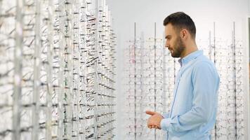 Side view of male customer choosing eyewear while standing near shelf during shopping at optical shop video