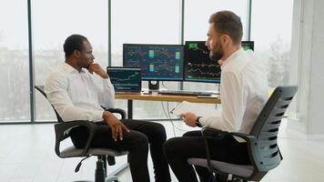 Two men traders sitting at desk at office together monitoring stocks data candle charts on screen video