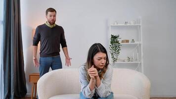 Hopeless young woman sitting on the sofa at home, while her husband abuser screaming at her, expressing aggression. Domestic violence and emotional abuse concept video