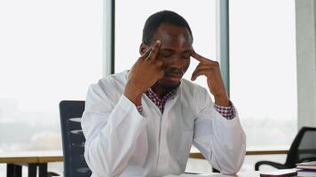 Stressed frustrated black doctor in white uniform with headache sitting alone at work desk in hospital. Sad young gp feeling desperate and guilty for medical failure video