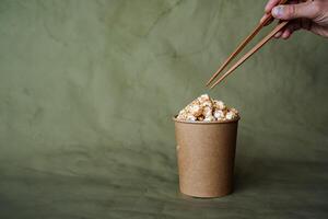 Chinese chopsticks to take popcorn from a box, a full can of popcorn, minimalistic style, on a colored background, concept picture, abstraction photo