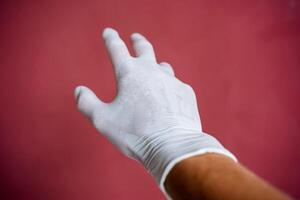 un mano en un blanco médico guante estiramientos adelante, un sudoroso mano debajo látex, dedos untado fuera en contra un granate muro, mano proteccion foto