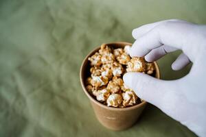 a hand takes sweet popcorn from a brown jar, a box of popcorn on a colored background, a gloved hand reaches for fast food, a minimalistic concept photo
