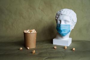 the concept of feeding a person in a medical mask with food, spilling food in front of the face in a mask, pouring popcorn from a jar on the table, plaster head in a protective mask, quarantine, art photo