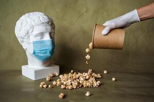 the concept of feeding a person in a medical mask with food, spilling food in front of the face in a mask, pouring popcorn from a jar on the table, plaster head in a protective mask, quarantine, art photo