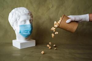 the concept of feeding a person in a medical mask with food, spilling food in front of the face in a mask, pouring popcorn from a jar on the table, plaster head in a protective mask, quarantine, art photo