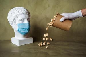 the concept of feeding a person in a medical mask with food, spilling food in front of the face in a mask, pouring popcorn from a jar on the table, plaster head in a protective mask, quarantine, art photo