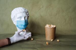 the concept of feeding a person in a medical mask with food, spilling food in front of the face in a mask, pouring popcorn from a jar on the table, plaster head in a protective mask, quarantine, art photo