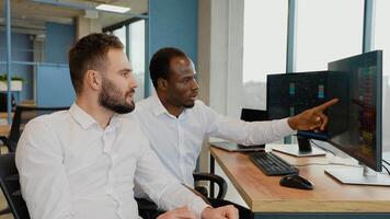 Two diverse colleagues traders talking to each other, looking at graphs while sitting in the office in front of multiple computer screens video